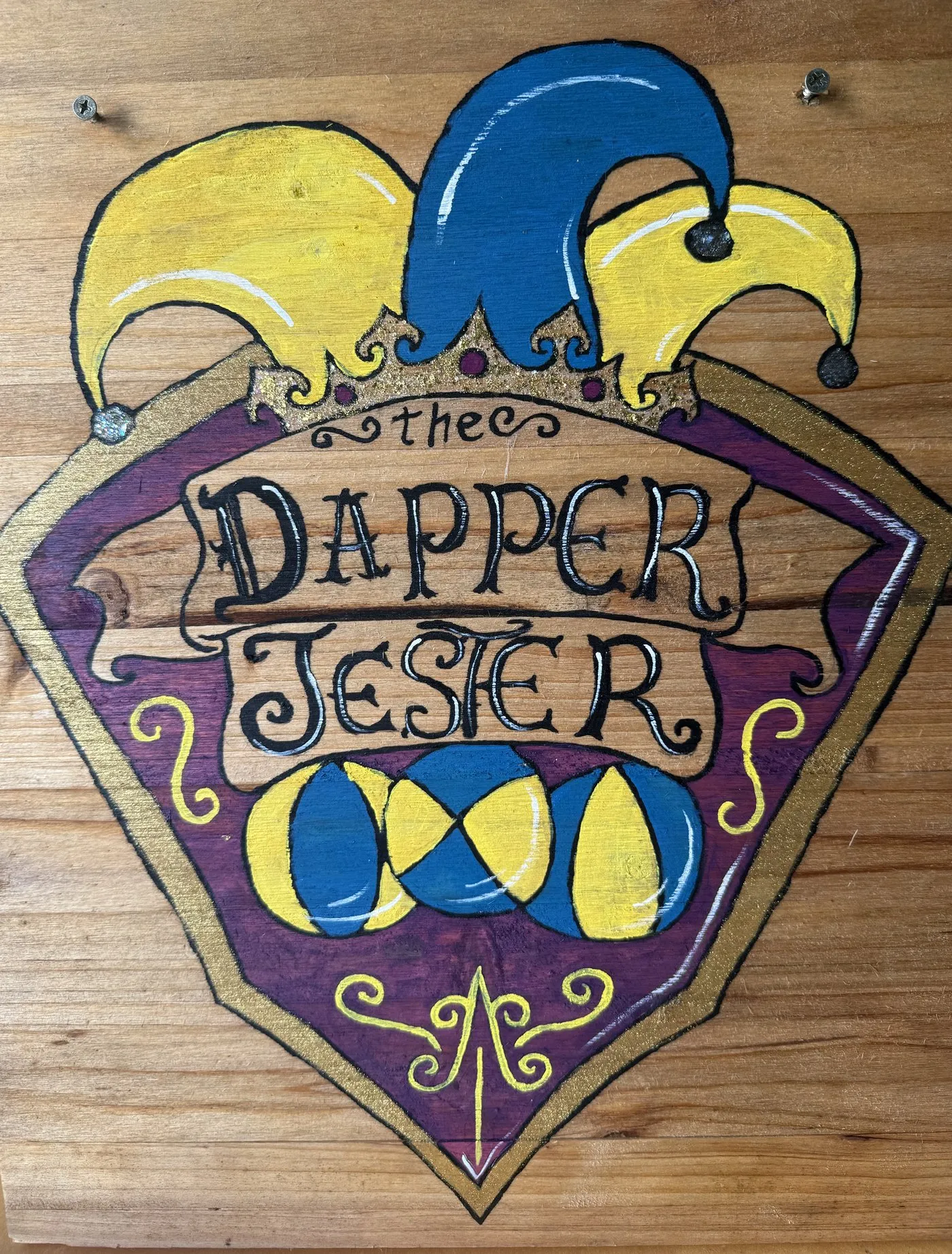 The Dapper Jester's hand-painted wooden booth sign: a plum crest topped with a blue-and-gold jester hat, over three juggling balls